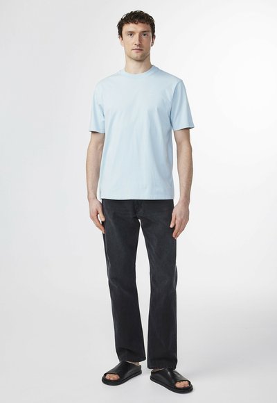 Camiseta de algodón azul claro con cuello redondo, combinada con jeans rectos negros y sandalias negras de tipo deslizante, de pie contra un fondo blanco.