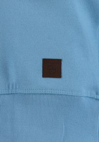 Sudadera azul claro con dobladillo acanalado y una etiqueta de cuero marrón con el texto "KEY LARGO". Textura suave con un diseño minimalista.
