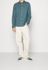 Camicia a maniche lunghe teal con bottoni, pantaloni larghi color crema e sneakers grigie con strisce bianche. Motivo semplice, colletto standard, vestibilità comoda.