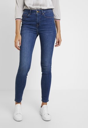 Högmidjade blå skinny jeans i denim, med framfickor, subtil sömnad och en slät yta, i sällskap med vita sneakers.