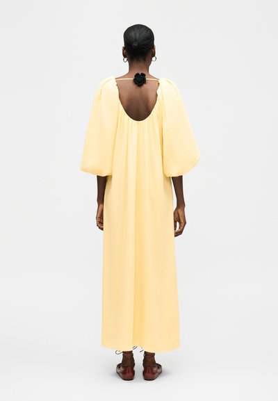 Γυναίκα με σκούρο δέρμα και μαλλιά σε κότσο φοράει ένα μακρύ ανοιχτό κίτρινο φόρεμα με φουσκωτά μανίκια και ανοιχτή πλάτη, στερεωμένο με μαύρη λεπτομέρεια σε σχήμα λουλουδιού.