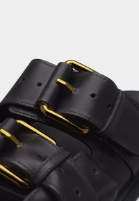 Des brides de sandale en cuir noir avec des boucles en métal doré et plusieurs trous d'ajustement, présentées en gros plan.