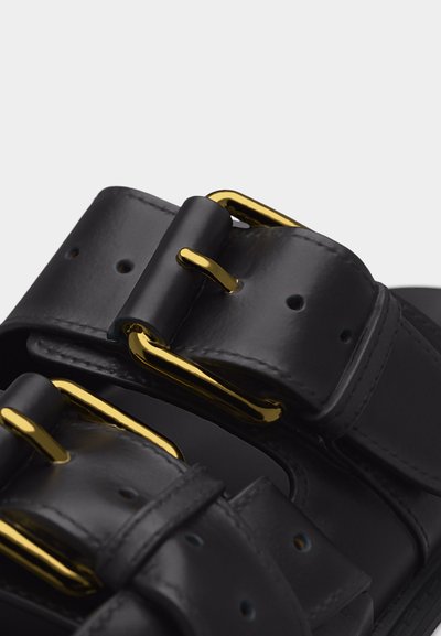 Λουριά μαύρου δερμάτινου σανδαλιού με αγκράφες από μέταλλο σε χρυσή απόχρωση και πολλαπλές τρύπες για ρύθμιση, εμφανιζόμενα σε κοντινό πλάνο.