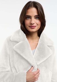 Jonge vrouw met schouderlang bruin haar, gekleed in een witte pluche jas, met een hand de kraag vasthoudend, en zachtjes lachend naar de camera.