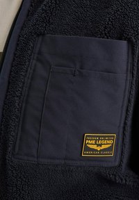 Detalj mornarskog flis jakne s teksturiranim materijalom, velikom preklopnom džepom i žutim izvezenim PME Legend logotipom.