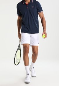 Muž oblečený v námořnickém polo, bílých šortkách a bílých teniskách Nike, držící tenisovou raketu a žlutý tenisový míček, jde vpřed.