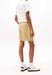 Mężczyzna w beżowych szortach z tylną kieszenią, białej koszuli z krótkim rękawem, białych skarpetkach i białych trampkach stoi na białym tle.