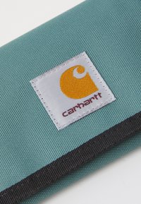 Billetera de la marca Carhartt en tela texturizada color teal, con un parche de logo en blanco y amarillo y ribete negro a lo largo del borde.