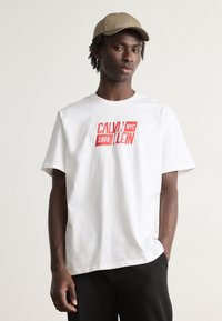 Bela bombažna majica s poudarjenim rdečim grafičnim tiskom "CALVIN KLEIN 1968 NYC" in kratkimi rokavi. Oblečena z črnimi hlačami in peščeno kapo.