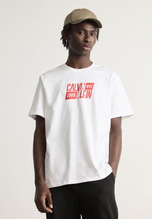 Bijela pamučna majica s izrazitim crvenim natpisom "CALVIN KLEIN 1968 NYC" i kratkim rukavima. Nosena je s crnim hlačama i bež kapom.