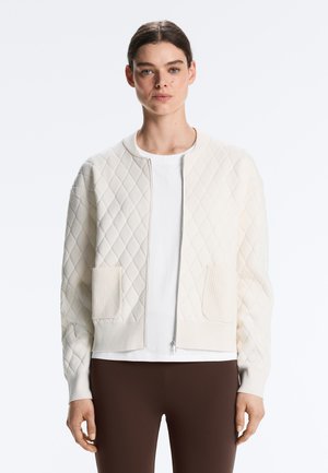 Jonge vrouw die een wit doorgestikt jasje met ritssluiting en twee voorzakken draagt, over een wit shirt en donkerbruine broek.