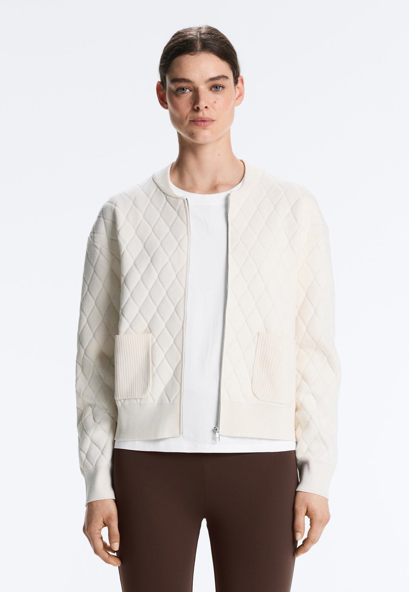 Jeune femme portant une veste blanche matelassée zippée avec deux poches avant, par-dessus une chemise blanche et un pantalon marron foncé.