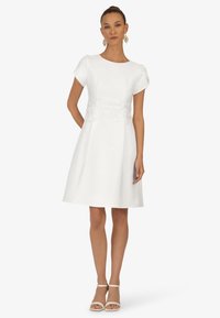 Robe blanche en ligne A avec de courtes manches bouffantes, détail en dentelle à la taille et texture de tissu lisse, associée à des talons blancs à bride de cheville.