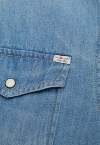 Jasnoniebieska tkanina dżinsowa z kieszenią, na której znajduje się mała biała metka z napisem "JACK & JONES SINCE 1990". Gładka tekstura, subtelne szwy.