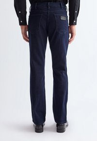 Vue de dos d'une personne portant un jean droit bleu foncé avec des poches arrière et des chaussures habillées noires sur un fond uni.