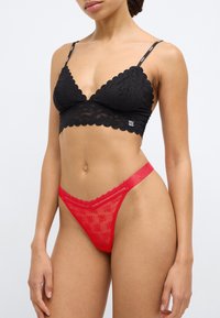 Bralette en dentelle noire avec bords festonnés et bretelles réglables, assortie à un string rouge transparent avec un motif logo et des détails festonnés.
