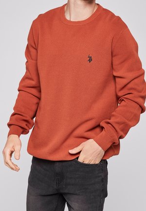 Pomarańczowy sweter o teksturowanej powierzchni, z okrągłym dekoltem, prążkowanymi mankietami i małym czarnym logo na piersi. Noszony z ciemnymi dżinsami.