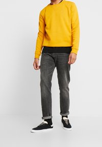 Sudadera amarilla con cuello redondo, parte inferior negra en capas, vaqueros grises y zapatillas negras con suela blanca. Textura suave, diseño casual.