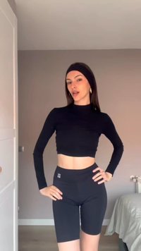 Femme debout à l'intérieur portant un crop top noir à manches longues, un short de cyclisme noir taille haute et un bandeau noir, posant les mains sur les hanches.