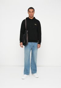 Hombre con sudadera negra, jeans azul claro, zapatillas blancas, y llevando una bolsa bandolera negra y gris, de pie contra un fondo blanco liso.