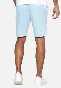 Personne portant un short bleu clair, des baskets blanches et une chemise blanche, se tenant de dos face à la caméra sur un fond uni.