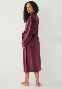 Vrouw met krullend haar in een lange, paarse fluwelen gewaad, vastgebonden in de taille, staande op blote voeten voor een effen witte achtergrond.