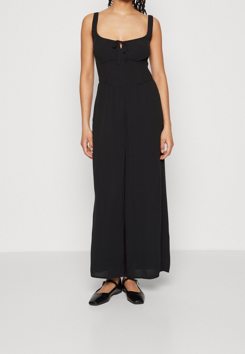 Kvinna iklädd en ärmlös svart jumpsuit med vida ben, fyrkantig halsringning och knytning vid bysten, matchad med svarta platta skor.