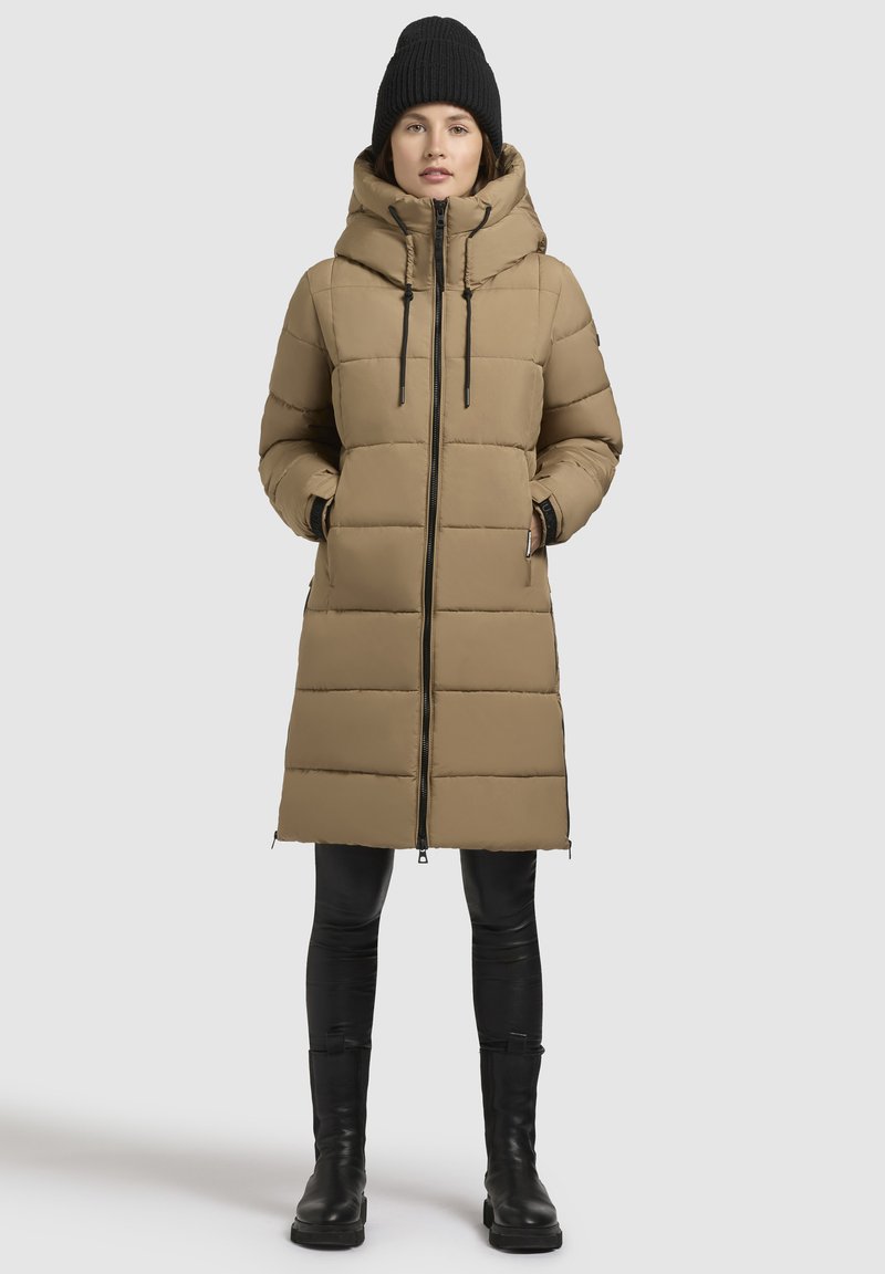 Beige puffer-takki, jossa on näkyvä huppu, vaakasuorat tilkkutäkkikappaleet ja edessä vetoketju. Yhdistetty mustiin housuihin ja korkeisiin saappaisiin.