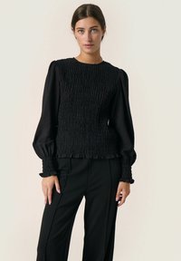 Black smocked top with long puff sleeves, textured fabric, and ruffled cuffs, paired with fitted black trousers. Simple design, no patterns.
