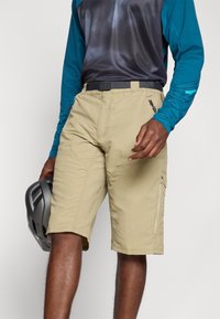 Shorts cargo beiges avec des poches zippées, conçus pour les activités de plein air, associés à un haut à manches longues bleu. Tenant un casque gris.