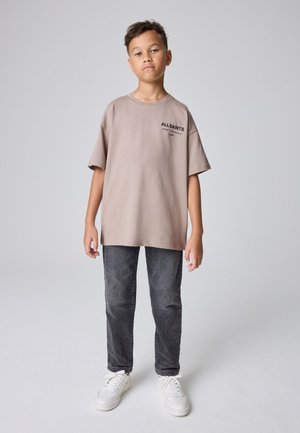 Chłopiec stoi, ubrany w beżową oversize koszulkę z napisem "ALLSAINTS", ciemnoszare dżinsy i białe sneakersy na jednolitym szarym tle.