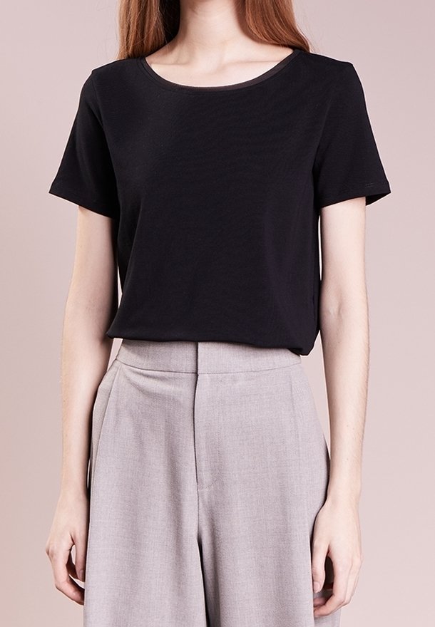 Femme portant un haut noir uni à manches courtes, rentré dans un pantalon large taille haute gris clair, sur un fond neutre.