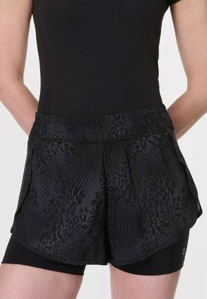 Femme portant un short de sport noir à motifs par-dessus un cuissard de cyclisme noir ajusté, debout avec les mains derrière le dos.