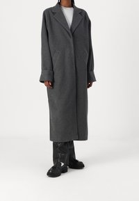 Lungo cappotto grigio in lana con colletto a labbra, tasche frontali e polsini larghi, indossato sopra una camicia grigia chiara e pantaloni neri slim.