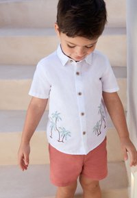 Chemise blanche à manches courtes avec broderie de palmier en vert et brun, boutons en bois, portée avec un short corail, debout sur des escaliers.