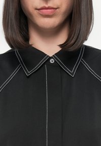 Black collared shirt with contrasting white stitching, featuring a button closure at the collar and a structured, tailored design.