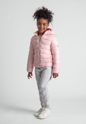 Glunderend kind staat rechtop, gekleed in een lichtroze gewatteerde jas met capuchon, grijze joggingbroek en witte sneakers op een effen achtergrond.