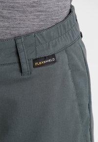 Harmaat, joustavaa kangasta olevat housut, joissa on joustava vyötärö. Housuissa on musta etiketti, jossa lukee "FLEXSHIELD" ja näkyvät ompelutiedot.