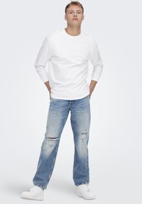 Maglietta bianca a maniche lunghe abbinata a jeans strappati blu chiaro e sneaker bianche, posa in piedi davanti a uno sfondo semplice.