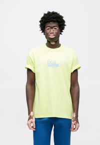 Žlté bavlnené tričko s rolovanými rukávmi, na prednej strane modré grafické logo „Café Swoosh“. Nosí sa s modrými nohavicami.