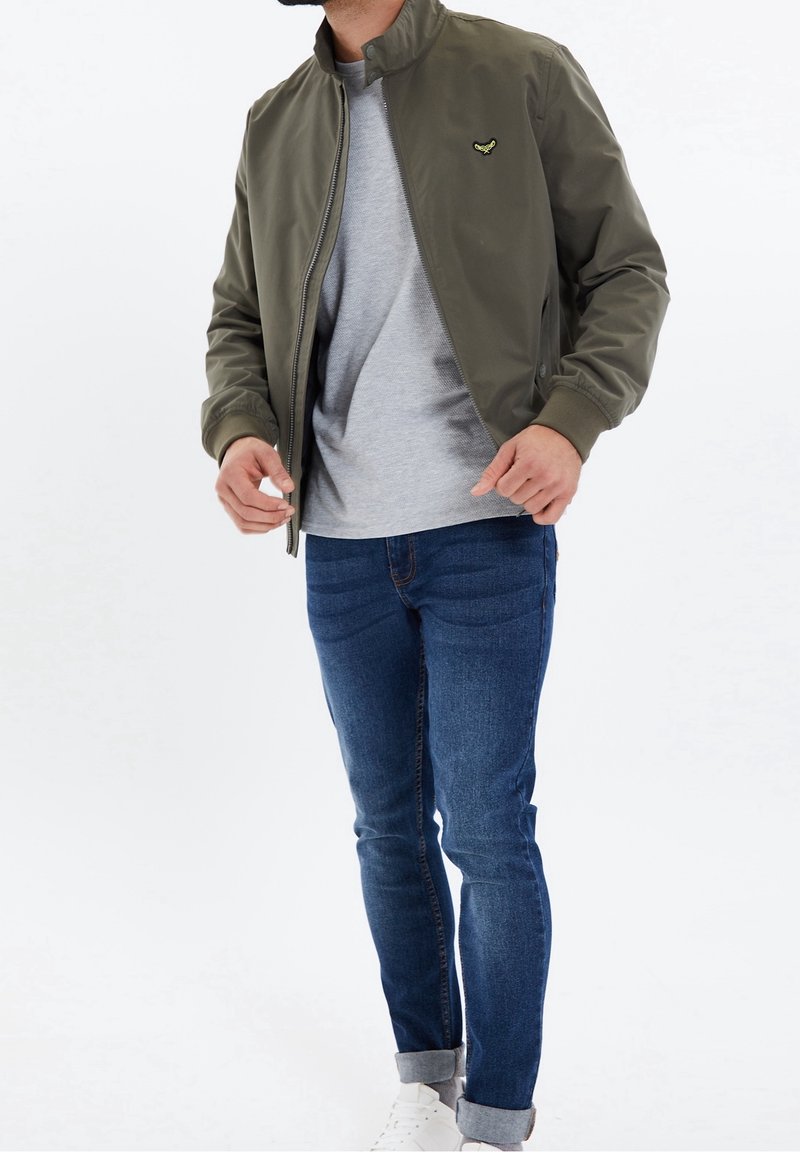 Homme portant une veste bomber vert olive sur une chemise grise, un jean bleu retroussé aux chevilles et des sneakers blanches, ajustant la fermeture éclair de la veste.
