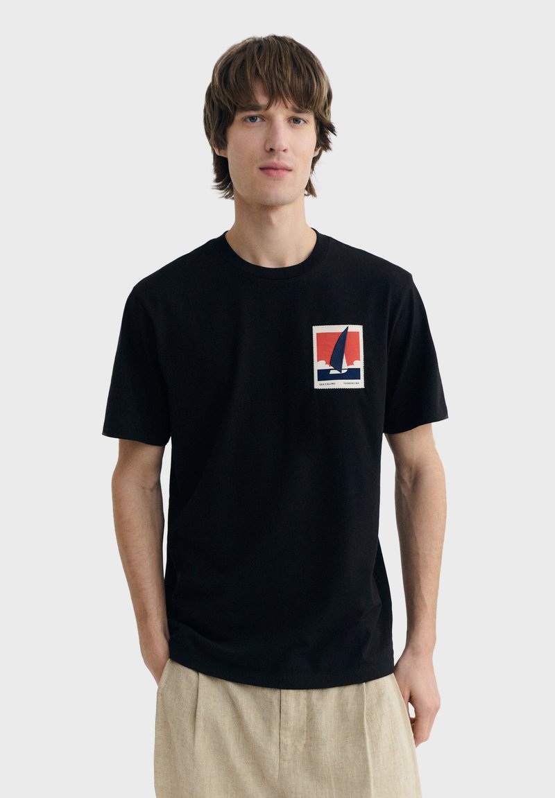 Jeune homme portant un t-shirt noir avec un graphisme de voilier rouge, blanc et bleu sur la poitrine, associé à un pantalon beige.