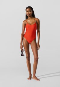 Rød badedragt i ét stykke med tynde stropper, en scoop-udskæring og minimal dækning. Modellen holder sorte solbriller og står foran en neutral baggrund.