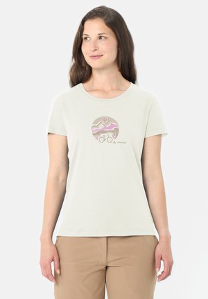 Donna che indossa una maglietta color beige chiaro con una grafica circolare di montagna, sole e bicicletta, abbinata a pantaloni color cuoio, in piedi su uno sfondo bianco.