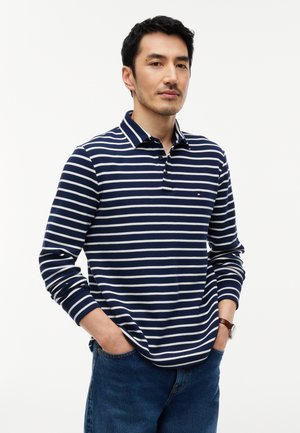 Homme portant un polo à manches longues rayé bleu marine et blanc avec col, debout les mains dans les poches devant un fond uni.