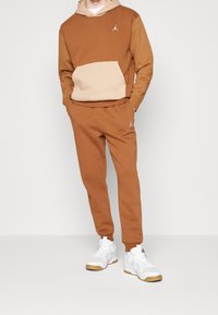 Felpa in cotone marrone e beige con tasca frontale, abbinata a pantaloni jogger marroni coordinati. Completata con un logo bianco e sneakers bianche.