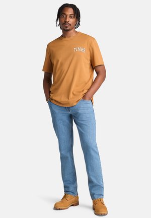 Braunes Baumwoll-T-Shirt mit "TIMBS" Grafik. Helle blaue Jeans, strukturierte beige Schuhe mit Schnürdesign, stehende Pose.