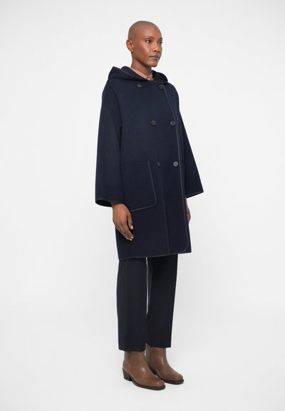 Palton din lână bleumarin cu glugă, cu nasturi dubli în față și buzunare mari. Asortat cu pantaloni negri croiți și botine maro cu toc.