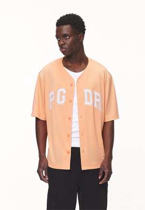 Kortærmet, ferskenfarvet skjorte med knapper og hvid tekst "PG DR." Har en afslappet pasform og let materiale. Sorte bukser.