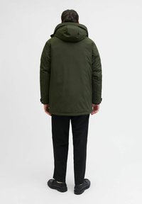 Donkergroene gewatteerde jas met capuchon, met een hoge kraag, een lichte glans en zwarte broek. Draag het met eenvoudige zwarte schoenen.